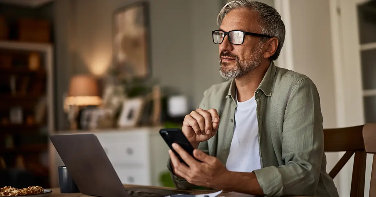 Man sitting at a table at home working on a laptop and using his mobile phone