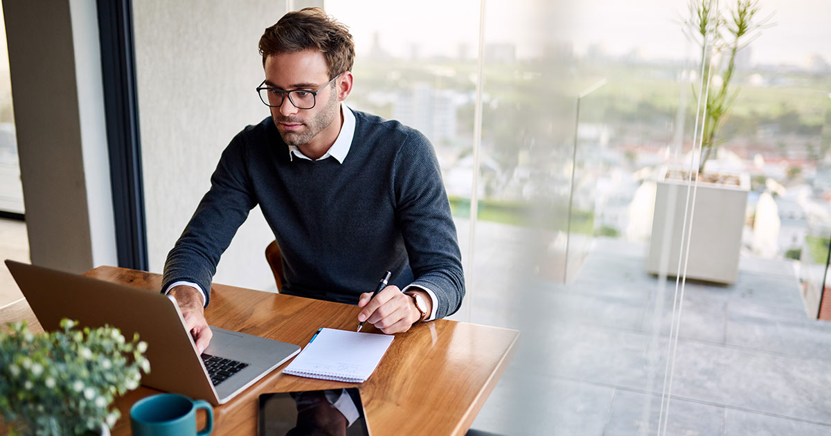 Man sitting at a table at home working on a laptop and writing down ideas in a notebook