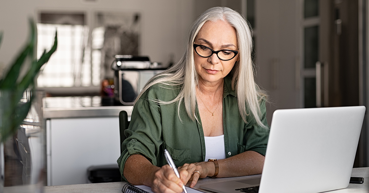 woman taking notes in notebook while using laptop at home