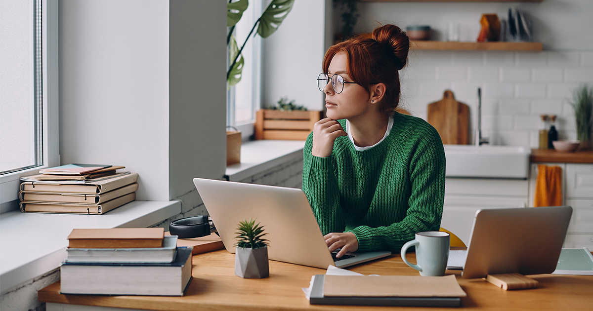 Red head woman wearing a green sweater looking thoughtful while working on laptop at the kitchen counter