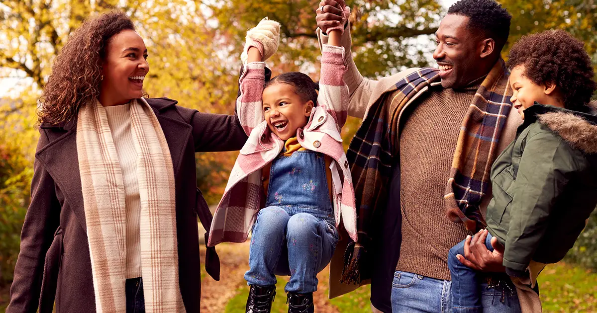 Smiling Family Having Fun Swinging Daughter On Walk Through Countryside Against Autumn Trees.