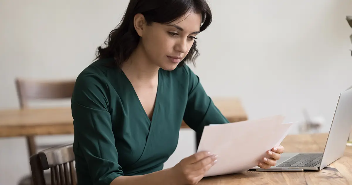 woman sitting at home office table with laptop reading accounting papers bills for payment