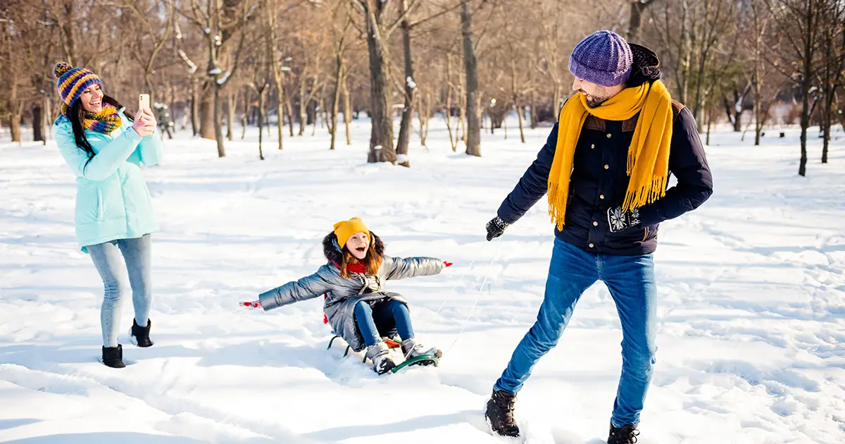 happy cheerful smiling family having good time outdoors little girl riding sled mother take photo on phone