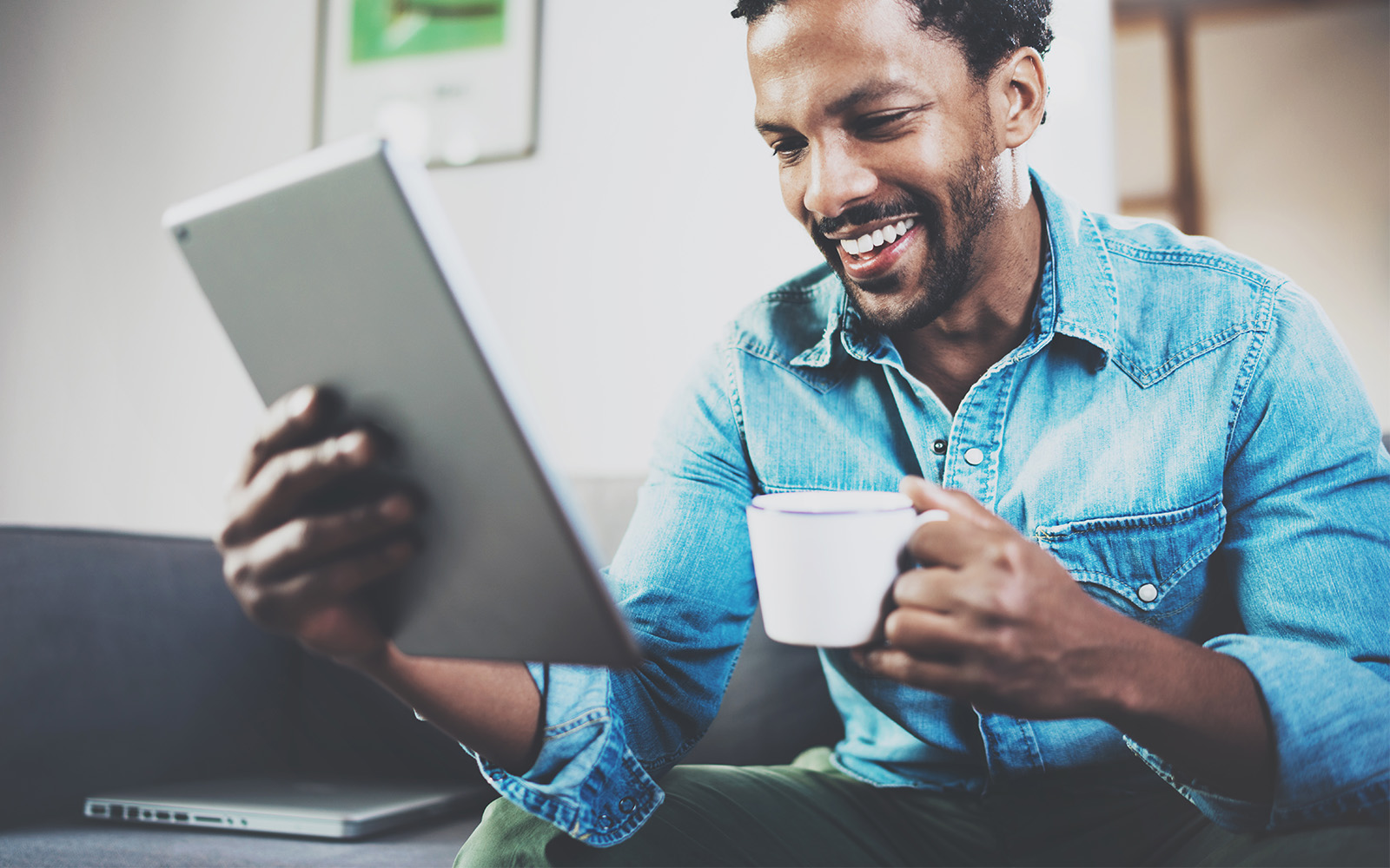 Person using tablet while sitting on sofa and holding white cup coffee in hand at home