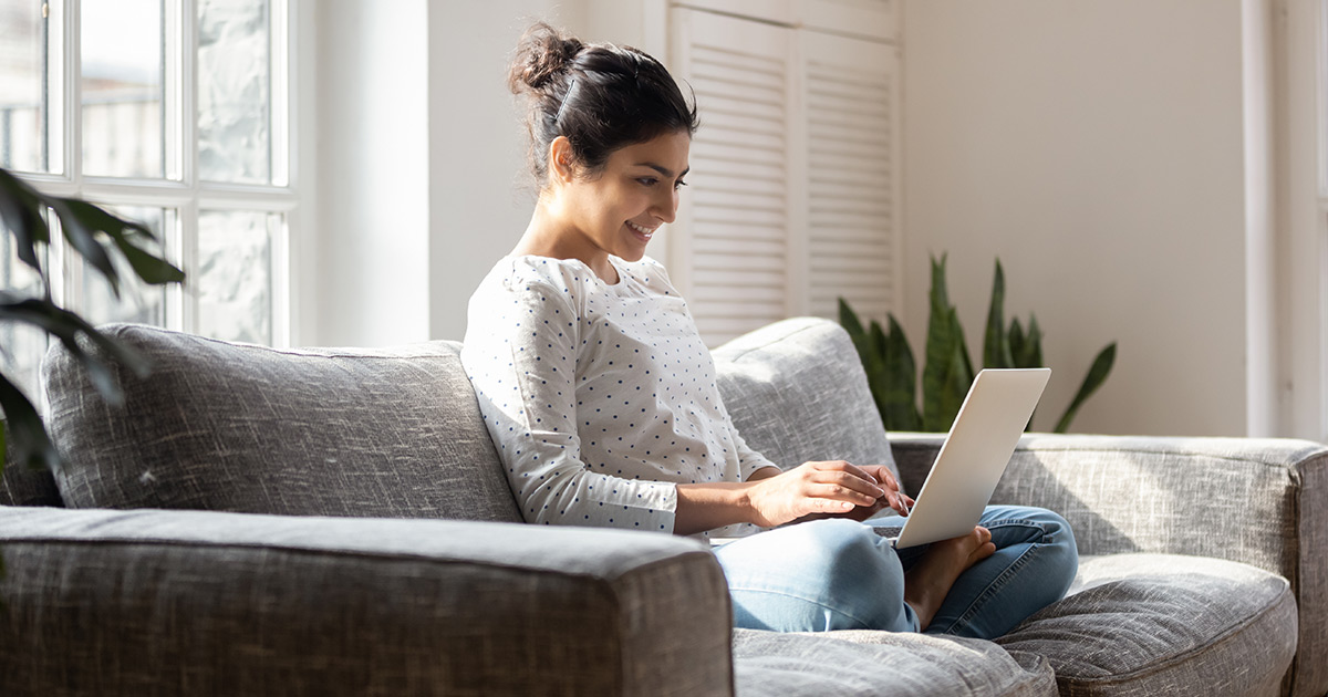 Woman sitting at her couch while typing on a laptop.